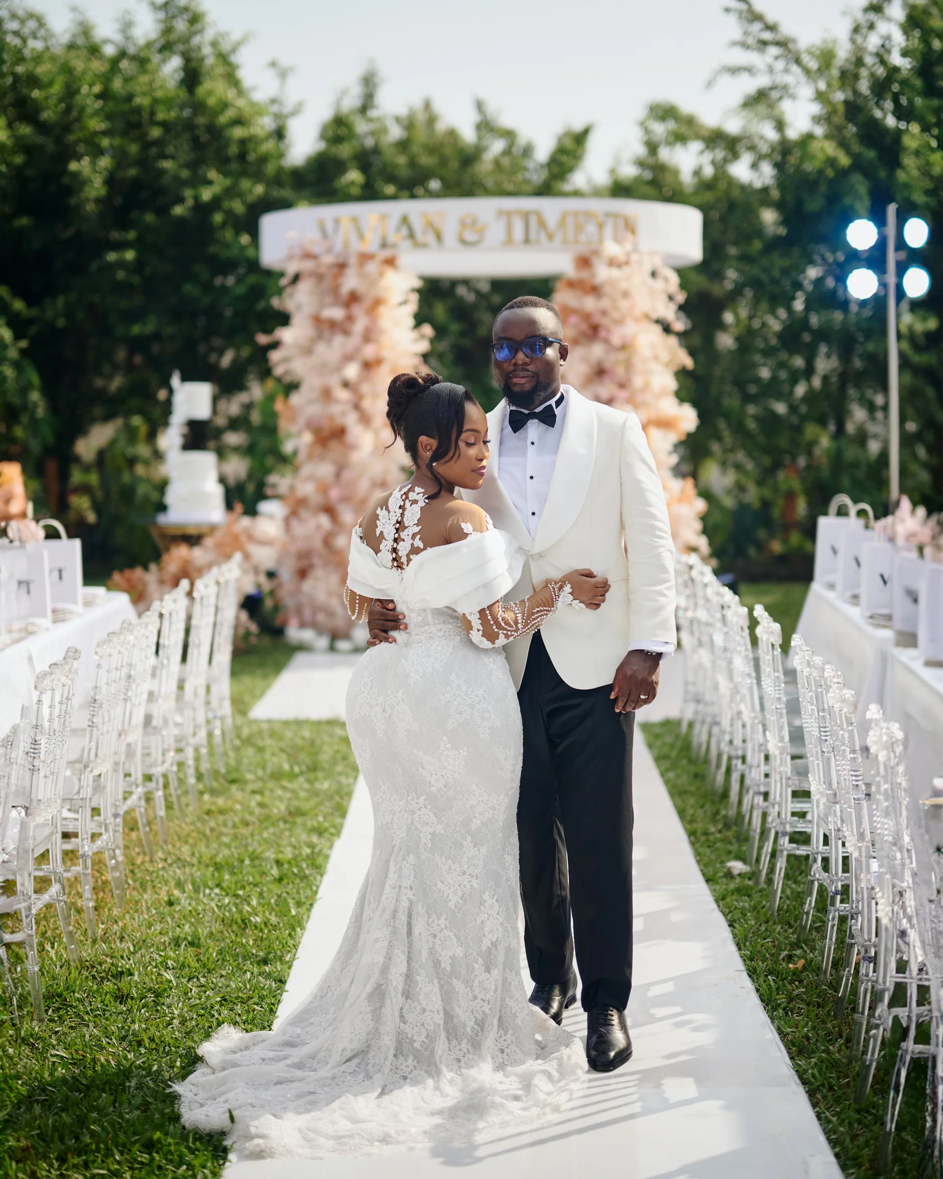 Bride and groom walking arm in arm past a floral arch reading "Vivian & Timeyin"