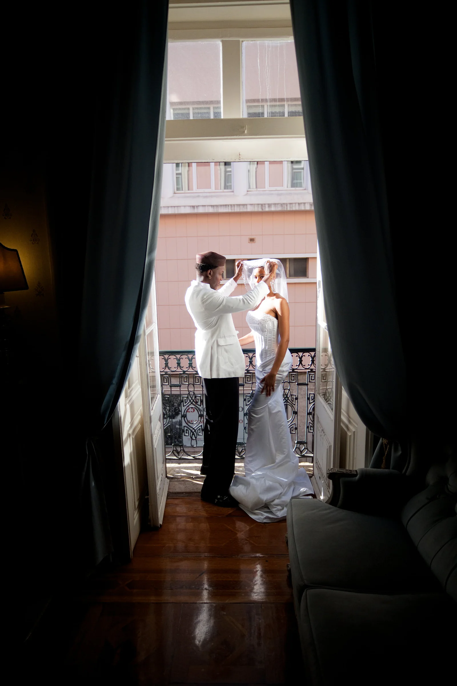 Couple on a wrought-iron balcony overlooking a pink European building facade