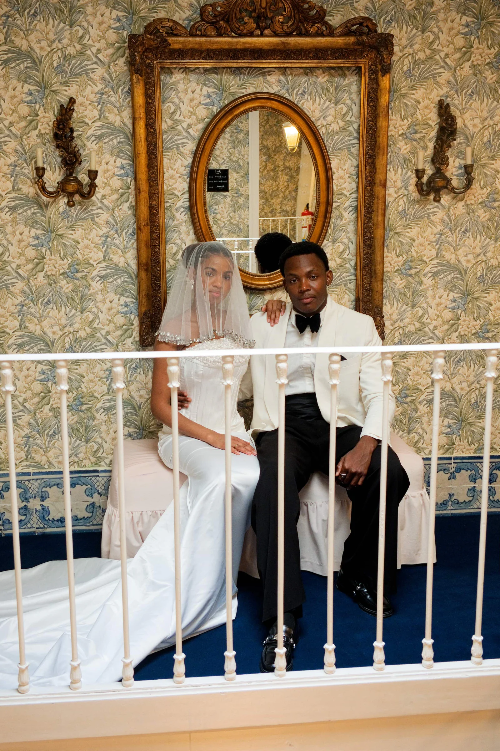 Bride and groom beside a four-poster bed in a room with floral wallpaper and blue tile