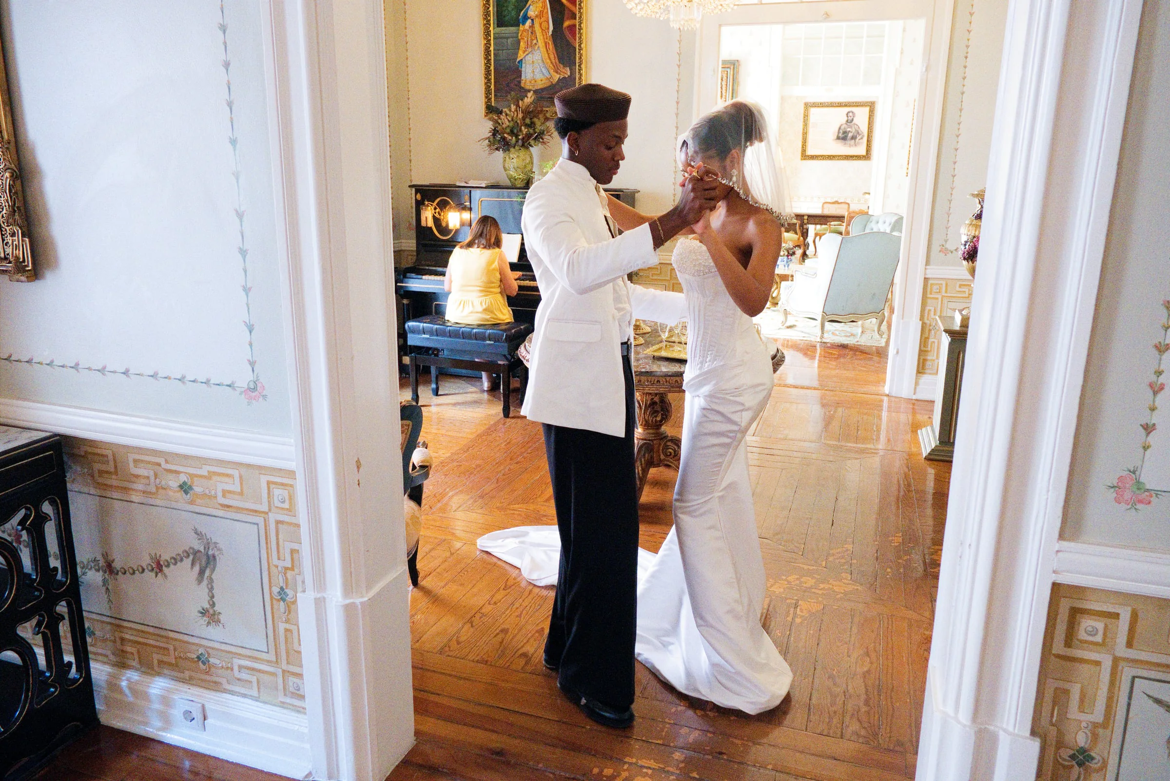 Groom adjusting his bride’s veil in a gilded manor hallway as a pianist plays