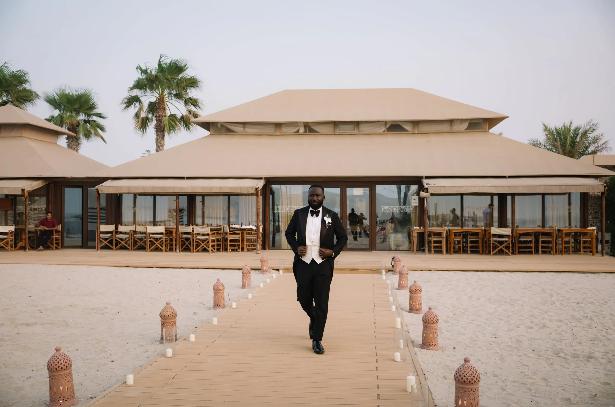 Groom in a tuxedo walking a candlelit boardwalk toward the venue