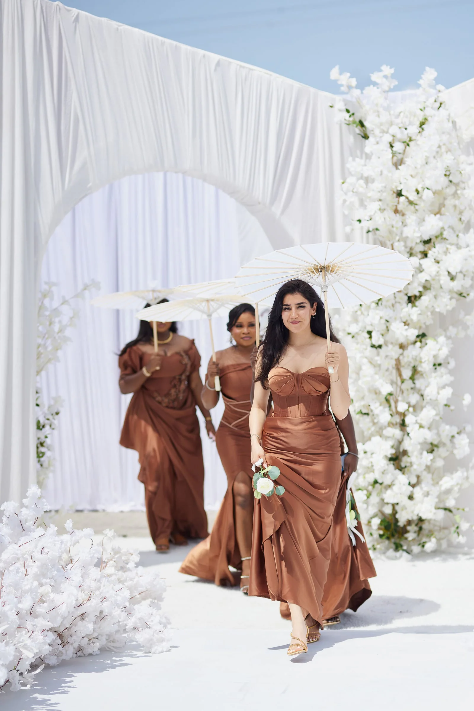 Bridesmaids in copper gowns carrying parasols beneath a white draped arch