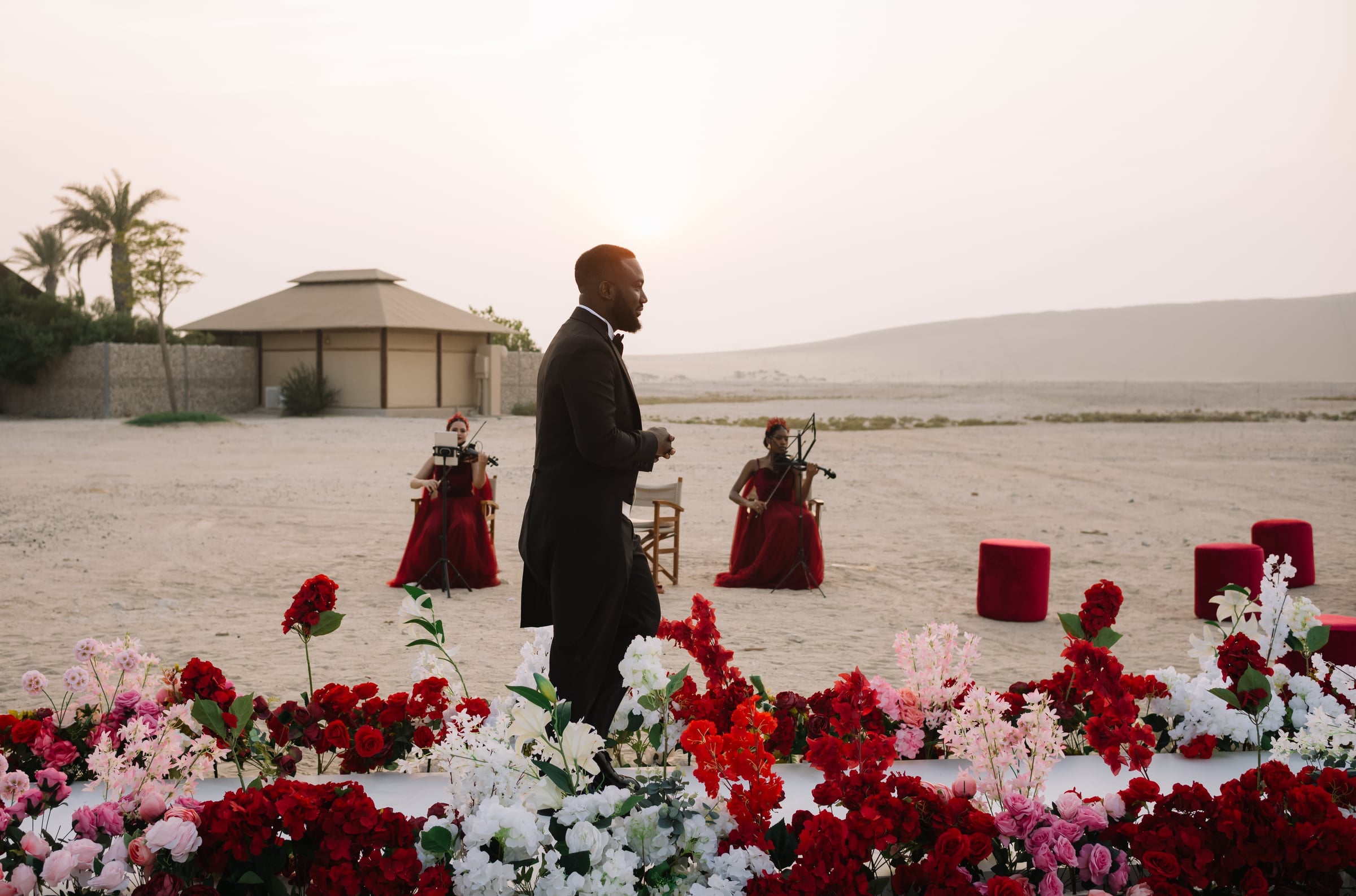Groom walking past violinists in red gowns on the sand at sunset